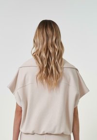 Haut ample couleur beige clair avec des manches larges et un ourlet arrondi. Tissu doux avec une texture lisse. Cheveux ondulés et lâches.