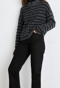 Cárdigan a rayas en carbón y negro con un cuello alto, que presenta un tejido suave y texturizado. Combinado con pantalones negros ajustados.