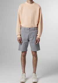 Felpa arancione chiaro con spalle cadute, pantaloni corti grigi e sneakers bianche. L'outfit presenta tessuti morbidi e un design casual.