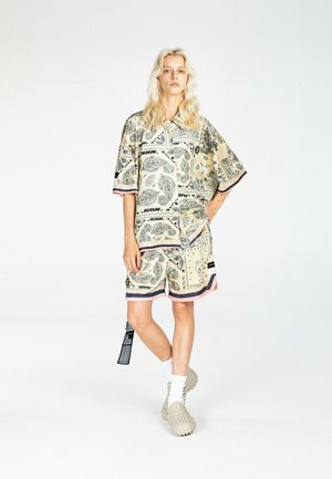 Persona rubia de pie con camisa grande beige con patrón de cachemira y pantalones cortos a juego, calcetines blancos y zapatos beige gruesos sobre fondo blanco.