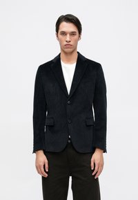 Blazer de veludo cotelê preto com gola lapela, dois bolsos frontais e fecho de um botão. Usado sobre uma t-shirt branca e calças escuras.