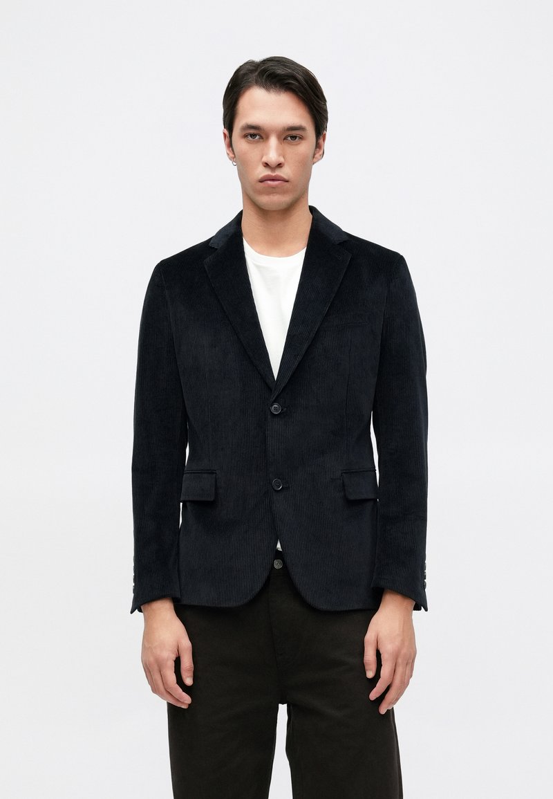 Blazer de veludo cotelê preto com gola lapela, dois bolsos frontais e fecho de um botão. Usado sobre uma t-shirt branca e calças escuras.