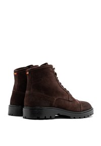 Brązowe zamszowe botki do kostki z okrągłym noskiem, czarną gumową podeszwą, metalowymi oczkami i detalem logo; posiadają małe kolorowe akcenty w postaci flag.