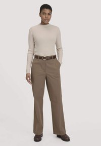 Rippens beiges Langarmshirt, kombiniert mit beigefarbenen Hosen und einem braunen Ledergürtel. Braune Schuhe runden das Outfit ab.