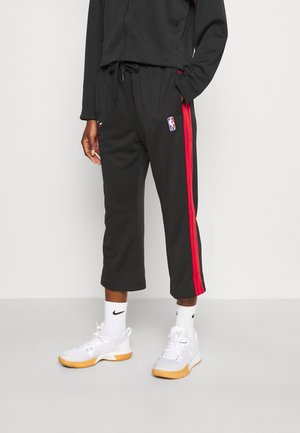 Pantalon de sport noir avec des bandes rouges sur les côtés, arborant un logo NBA. Associé à des baskets blanches et des chaussettes mi-mollet. Tissu doux, design court.
