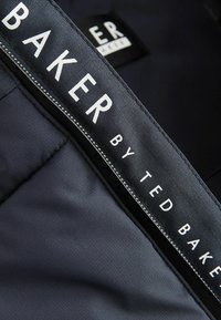 Étiquette en tissu marine avec "BAKER" et "BY TED BAKER" en texte blanc, brodée avec un bord contrastant, sur un fond texturé lisse.
