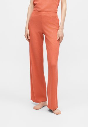Personne debout pieds nus portant un pantalon en maille côtelée corail ample et un haut assorti sur un fond uni.