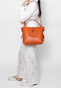 Borsa a secchiello in pelle arancione con parte superiore piatta e hardware in argento. Dispone di una tracolla removibile e superficie testurizzata.