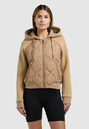 Femeie care poartă o hanorac căptușit bej cu fermoar și șnururi roșii, și pantaloni scurți negri de biciclist, stând în picioare pe un fundal simplu, deschis la culoare.