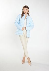 Giacca puffer blu chiaro con colletto rialzato e cuciture orizzontali, indossata sopra una maglietta bianca e pantaloni beige, completata da tacchi nudi.