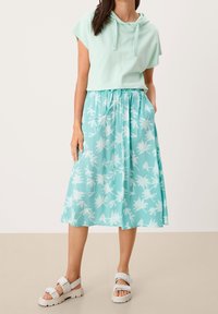 Hoodie vert clair court avec cordon de serrage. Jupe midi fluide couleur menthe avec imprimé de feuilles de palmier blanches et poches. Sandales blanches.