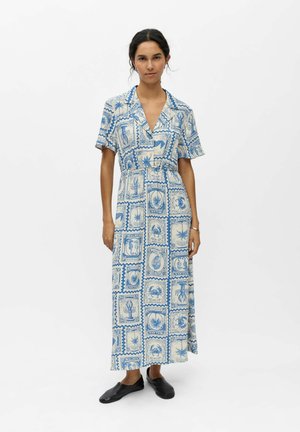 Femme portant une robe longue à manches courtes patronnée en bleu et blanc avec un col, debout devant un fond blanc uni.