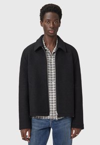 Zwarte wolmix jas met een kraag, ritsluiting en zijvakken, gelaagd over een grijs geruit overhemd met knopen, gecombineerd met blauwe jeans.