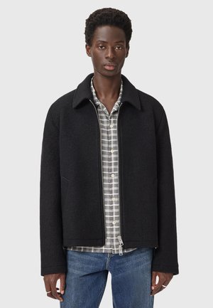 Veste en mélange de laine noire avec un col, fermeture éclair et poches latérales, superposée à une chemise à carreaux grise à boutons, associée à un jean bleu.