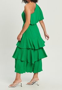 Zelené šaty na jedno rameno s vrstvenými volány, splývavým designem a lehkým materiálem. Nosené s bílými sandály na podpatku.