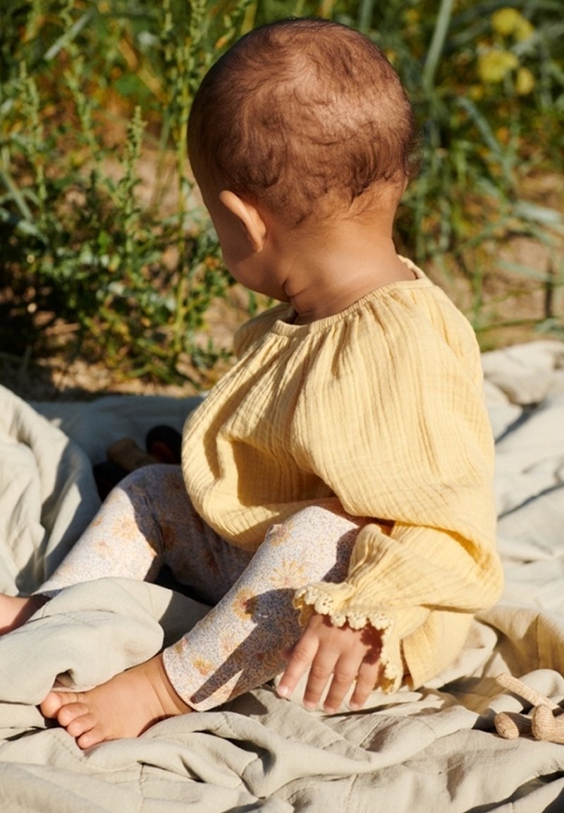 Bébé portant un chemisier jaune à manches longues et un legging à motifs floraux, assis sur une couverture en plein air avec des plantes vertes en arrière-plan.