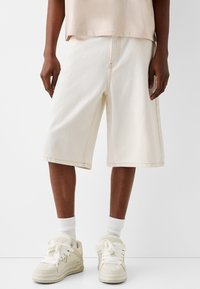 Kermaväriset farkkushortsit, joissa on sivutaskut, näkyvät ompeleet ja rento istuvuus, yhdistettyinä valkoisiin tennareihin ja valkoisiin sukkiin.