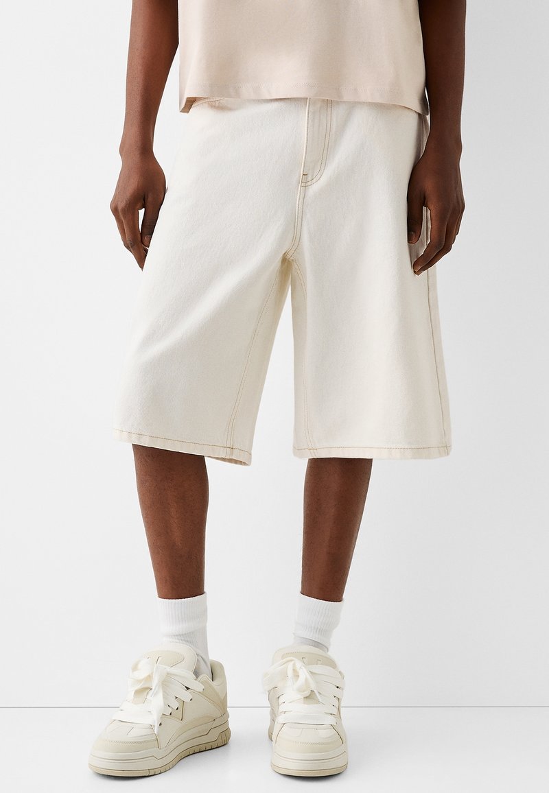 Kermaväriset farkkushortsit, joissa on sivutaskut, näkyvät ompeleet ja rento istuvuus, yhdistettyinä valkoisiin tennareihin ja valkoisiin sukkiin.