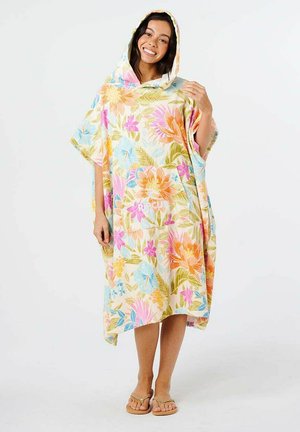 Woman wearing a hooded floral beach towel poncho with pastel orange, pink, blue, and green flowers, smiling and standing barefoot.