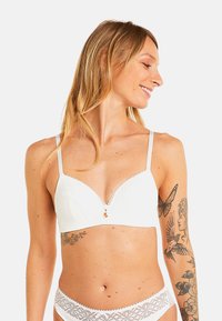 Bralette en dentelle blanche avec un décolleté en V et des bretelles réglables, ornée de motifs floraux et d'un petit accent décoratif au centre.