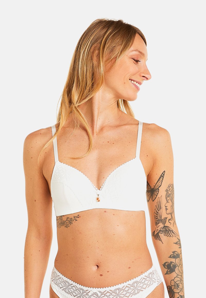 Bralette en dentelle blanche avec un décolleté en V et des bretelles réglables, ornée de motifs floraux et d'un petit accent décoratif au centre.