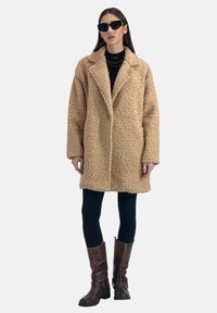 Cappotto in pelliccia sintetica beige con finitura testurizzata, forma oversize e rever a pistola. Indossato sopra un dolcevita nero e abbinato a stivali marroni.