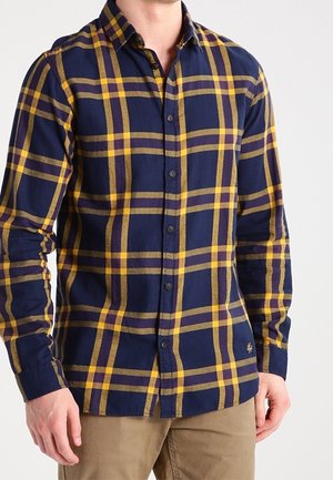 Mężczyzna w granatowo-żółtej kraciastej koszuli z długim rękawem na guziki i beżowych spodniach, stoi na jednolitym tle.