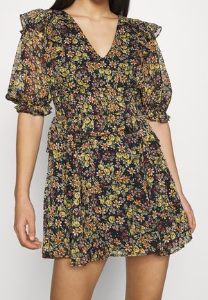 Kobieta w krótkiej czarnej sukience w kwiaty z bufiastymi rękawami, falbanowym dołem i dekoltem w kształcie litery V.