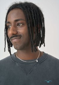 Schwarzer Sweatshirt mit einer subtilen Struktur, ausgestattet mit einem hellblauen Logo. Die Person hat geflochtenes Haar und trägt eine mehrlagige silberne Halskette.