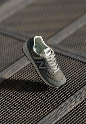 Szaro-biały but sportowy New Balance z sznurówkami, położony na teksturowanej metalowej siatce, oświetlony miękkim światłem.