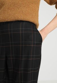 Černé kostkované kalhoty s hnědými a šedými pruhy, s hladkou texturou, boční kapsou a volným střihem. Nošeny s hnědým pleteným svetrem.