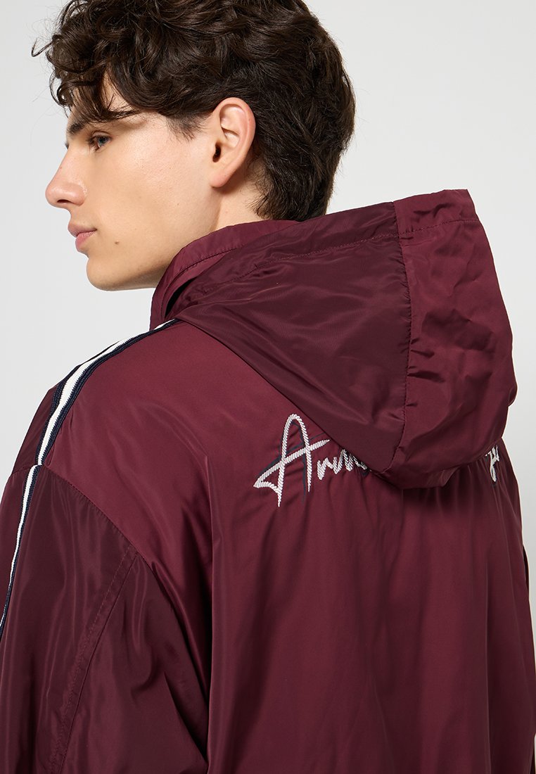 Giacca leggera bordeaux con cappuccio, caratterizzata da strisce bianche e navy lungo le maniche e testo ricamato sul retro in filo argentato.