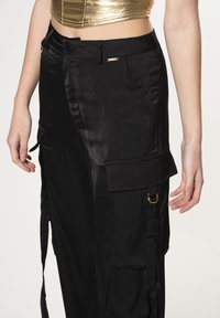 Zwarte cargobroek van satijn, met een zijvak, verstelbare bandjes en een kleine metalen accent op de tailleband. Gladde textuur.