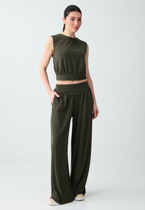 Kobieta ubrana w ciemnozielony, bezrękawnik typu crop top i szerokie spodnie z wysokim stanem, stojąca z jedną ręką w kieszeni, na jasnym tle.