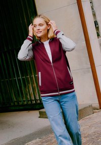 Vinrød jakke med lynlås, grå ærmer og hvide detaljer, med sidelommer og ribstrikkede manchetter. Kombineret med lyseblå jeans.