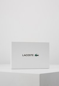 Λευκό καφέ με υφή με το "LACOSTE" τυπωμένο σε μαύρο, διαθέτοντας ένα πράσινο λογότυπο κροκόδειλου στη δεξιά πλευρά. Απλή, ορθογώνια σχεδίαση.
