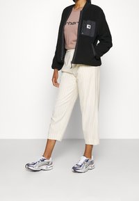 Czarna polarowa kurtka bombowiec z kieszenią na zamek, bladoróżowa koszulka z logo, kremowe wąskie spodnie w paski, oraz wielokolorowe sneakersy.