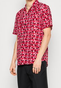 Koszula z krótkim rękawem w kolorze czerwonym z wyraźnym czarno-białym powtarzającym się wzorem tekstowym. Posiada kołnierz typu camp i luźny krój. Wykonana z lekkiego materiału.