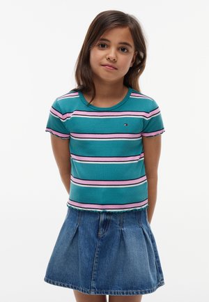 Junges Mädchen trägt ein kurzärmeliges, gestreiftes Shirt in Türkis, Pink und Weiß sowie einen blauen Jeansrock und steht mit den Händen hinter dem Rücken.
