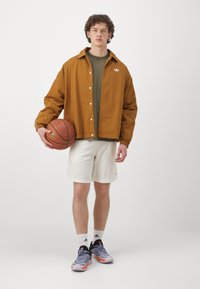 Jachetă maronie, cu nasturi, cu manșete elastice, cămașă verde dedesubt, pantaloni scurți albi, teneși gri, ținând o minge de baschet. Design simplu, croi lejer.