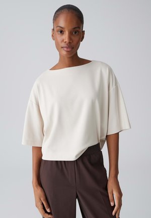 Frau mit dunkler Haut, die ein lockeres, weißes Kurzarmoberteil und dunkelbraune Hose trägt und vor einem einfachen hellgrauen Hintergrund steht.