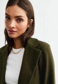 Olivgrön yllekappa med skarpa slag, bärs över en vit topp. En anmärkningsvärd guldchainhalsband framhäver designen.