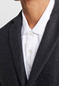 Grå uld blazer med knaphulslapper over en hvid skjorte med knapper. Stoffets tekstur er glat, med synlige knapper og syninger.