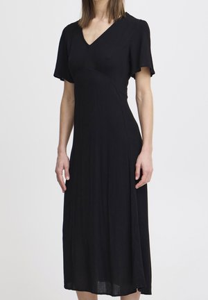 Kobieta w czarnej, krótkorękawowej sukience midi z dekoltem w serek, dopasowaną w talii i rozkloszowanym dołem, na jasnym, jednolitym tle.