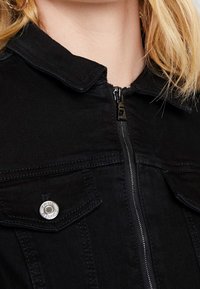 Black denim jacket featuring a zip front, pointed collar, and two breast pockets with silver snap closures. Textured fabric provides structure.