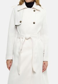 Manteau long en laine blanche avec un devant croisé, un grand col et une ceinture à nouer. Comprend des poches latérales et des boutons dorés.