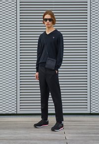 Svart v-ringad tröja, svarta byxor och svarta sneakers med röda accenter. Justerbar axelremsväska. Modellen står mot en texturerad bakgrund.