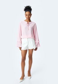 Blusa lunga rosa chiaro con maniche lunghe, scollatura tonda e dettagli con fiocco, abbinata a pantaloni corti bianchi sartoriali e tacchi bianchi a punta.