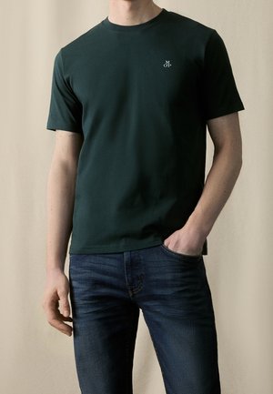Man wearing dark green short-sleeve t-shirt with small white "M OP" logo on chest, paired with dark blue jeans, hand in pocket.