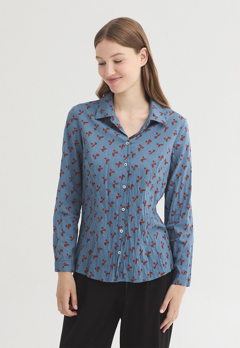 Jeune femme portant une chemise bleue boutonnée avec des motifs floraux rouges et un pantalon noir, regardant vers le bas et souriant doucement.